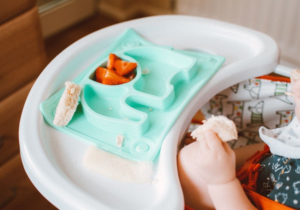 Baby in a high chair with an orange harness, white tray and mint elephant suction plate holding bread and strawberries