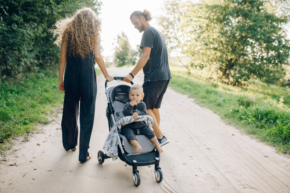 Parents pushing a light grey stroller with a toddler on a sunny park path